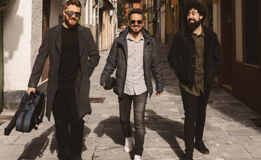 Kuvassa on kolme nuorta miestä kävelemässä kapeaa talojen reunustamaa katua. Heillä n mustat vaatteet paitsi keskimmäisellä harmaa paita.