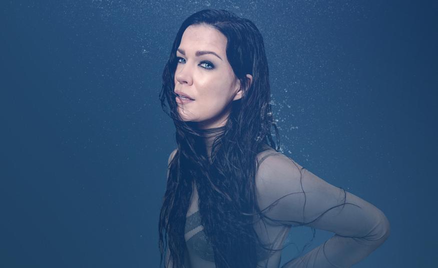 Kuvassa on sivuttain Jenni Vartiainen ja hänellä on tummat pitkät hiukset sekä hihoista läpinäkyvä pusero. Tausta on tumman sininen.