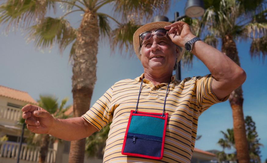 Kuvassa on Tom Lindholm kuvattuna yläviistoon. Hänellä on karttalaukku kaulassaan ja hän pitää aurinkolaseista kiinni vasemmalla kädellään. Taustalla on palmuja, vaaleita rakennuksia ja sinistä taivasta.