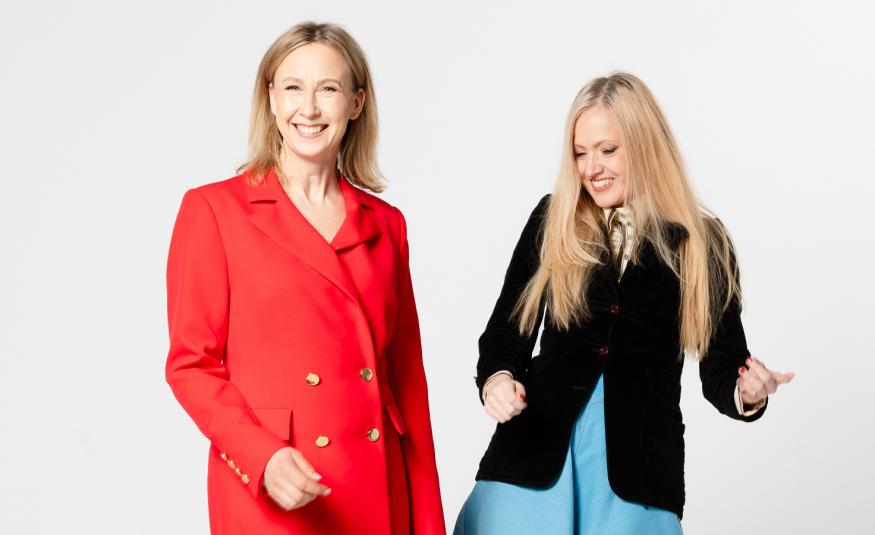 Kuvassa on vasemmalla punaisessa takkipuvussa Heli Meklin ja oikealla on Eveliina Talvitie jammailemassa mustassa jakussa ja vaalean sinisessä mekossa. Tausta on vaalea.