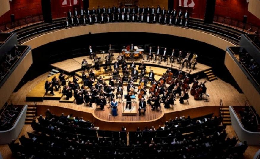Kuvassa on Sinfonia Lahti ylhäältä kuvattuna soikealla puisella esiintymislavalla soittamassa. Ylempänä näkyy parvekkeella yleisöä ja edessä permannolla. Kuva on osa kirjan kannesta.