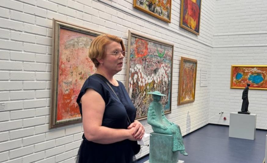 Kuvassa on Maarit Lavas sivuttain puhumassa tummassa mekossa. Taustalla on valkoisella tiiliseinällä värikkäitä teoksia kahdessa rivissä.