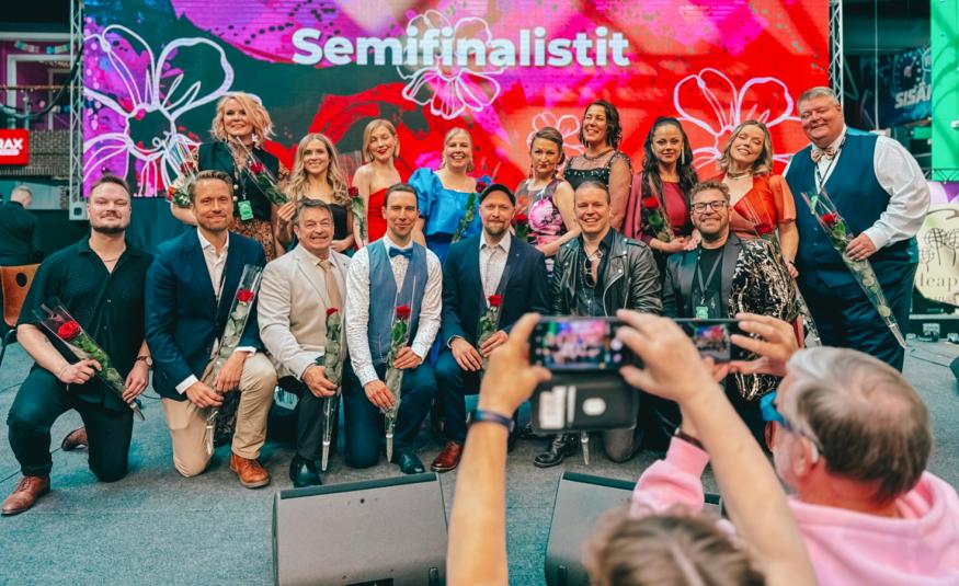 Kuvassa ovat kahdessa rivissä Tangomarkkinoiden semifinalistit ja heistä ensimmäisessä rivissä olevat nojaavat toiseen polveensa. Taustalla on punaisia kukkia Tangomarkkinoiden isossa lakanassa. Edessä on yleisöä ottamassa valokuvia.
