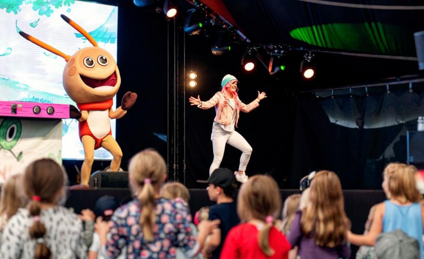 Kuvassa on esiintymislavalla vasemmalla Kikattava Kakkiainen, joka on oranssinen otus isoine päineen ja tuntosarvineen. Silla on punainen paita päällään. Lavalla oikealla on naislaulaja, jolla vaaleat vaatteet ja hän tekee loikkaa kädet levitettyinä.