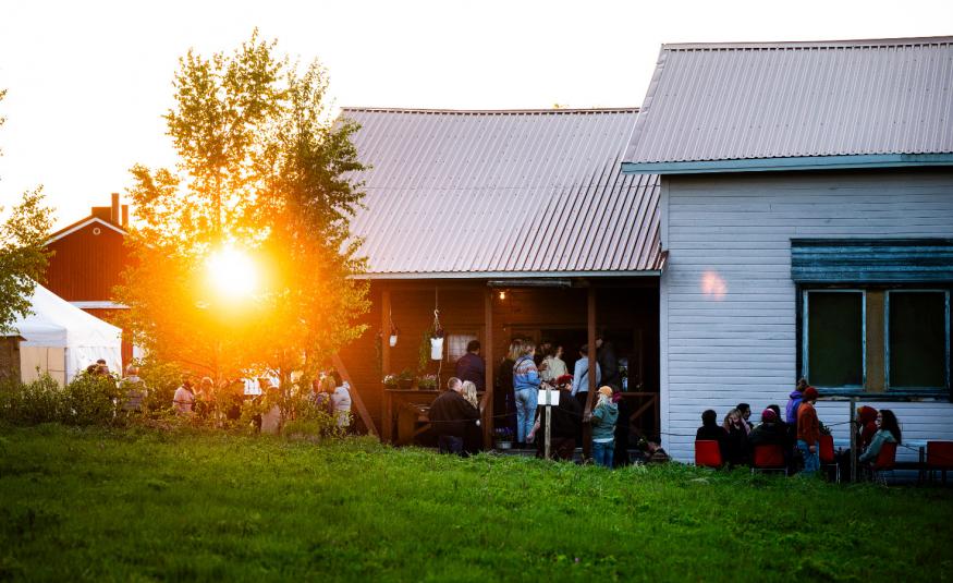 Kuvassa on valkoinen talo, jonka päässä on latomainen ja sen takaa paistaa ilta-aurinko. Rakennuksen edessä on ihmisiä ja vihreää nurmikkoa. 