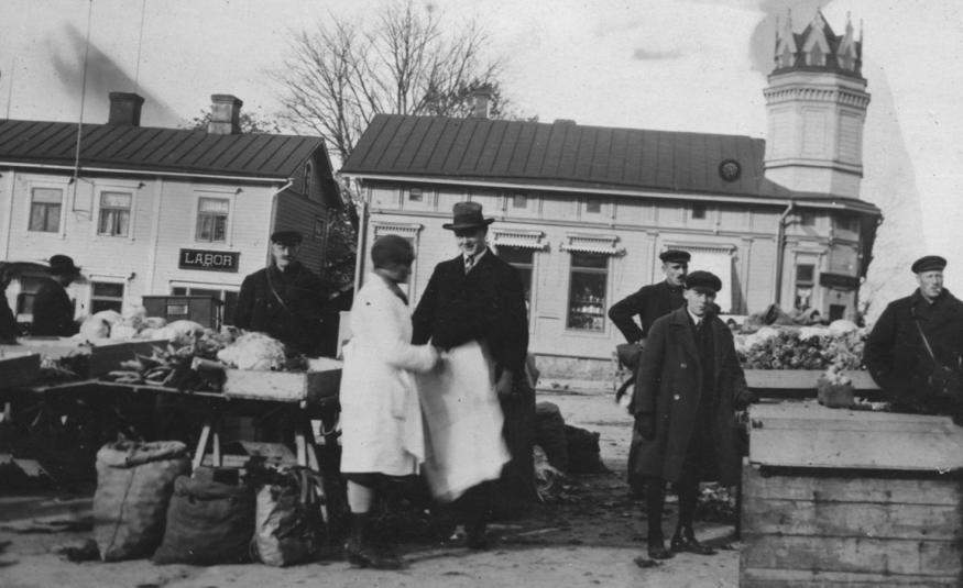 Kuvassa on etualalla torin myyntipöytä ja pöydän takana mies seisomassa. Pöydän vieressä seisoo nainen valkoisessa takissa ja mies mustassa hatussa ja lierihatussa. Oikealla on kaksi miestä ja välissä iso puulaatikko sekä sen vieressä mies. Taustalla on kaksi vaaleata, vanhaa puutaloa. Kuva on musta-valkoinen.