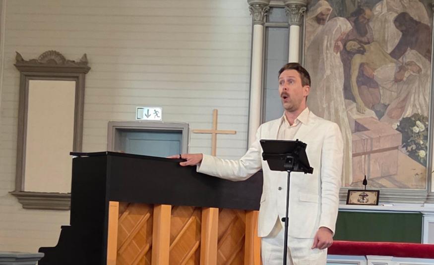 Kuvassa on Waltteri Torikka laulamassa oikealla valkoisessa puvussa ja hän pitää oikealla kättään pianon päällä. Pianosta näkyy ruskea, puinen takaosa
