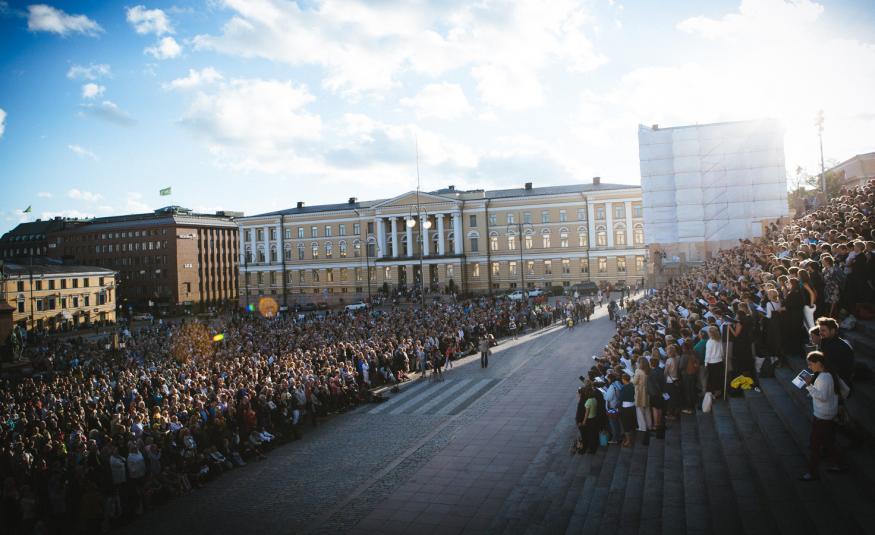 Kuvassa on Senaatintorilla vasemmalla runsaasti kuorolaisia seisomassa ja laulamassa. Oikealla on Helsingin tuomiokirkon portailla on myös runsaasti laulajia seisomassa. Keskellä on näkyvissä tilaa ja katukivetystä.