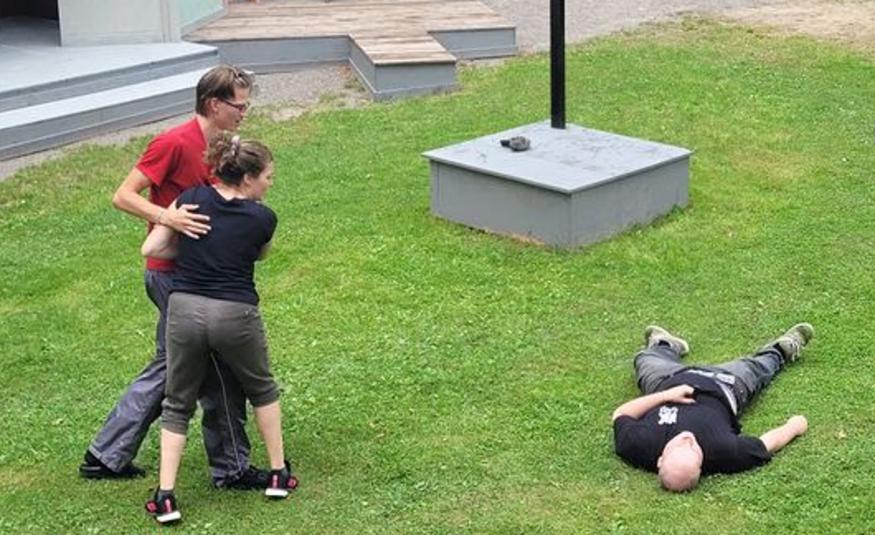 Kuvassa ovat vasemmalla nuori nainen ja mies ja he ovat jonkinlaisessa kamppailuotteessa ja oikealla katsovat maassa ruohikolla makaavaa nuorta miestä. Taustalla näkyy harmaita lavoja.