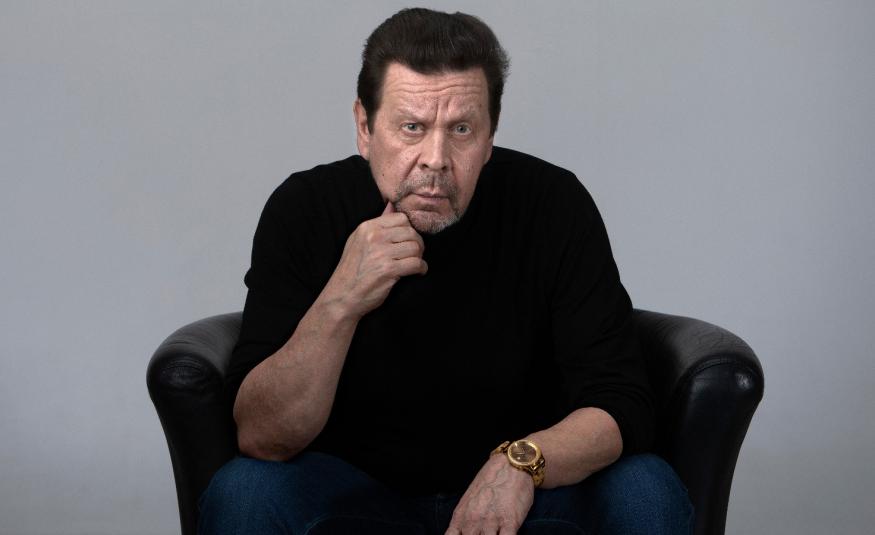 Kuvassa on Pate Mustajärvi mustassa paidassa istumassa mustassa isossa nojatuolissa kumartuneena eteenpäin. Hän nojaa oikealla kädellään leukaansa. Tausta on harmaa.