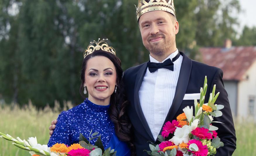 Kuvassa on vasemmalla Raija Mäntyniemi sinisessä mekossa ja vieressä oikealla Juho Punkeri mustassa takissa ja valkoisessa paidassa. Molemmilla on kukkakimput kädessään. Taustalla on metsää ja valkoinen talo.