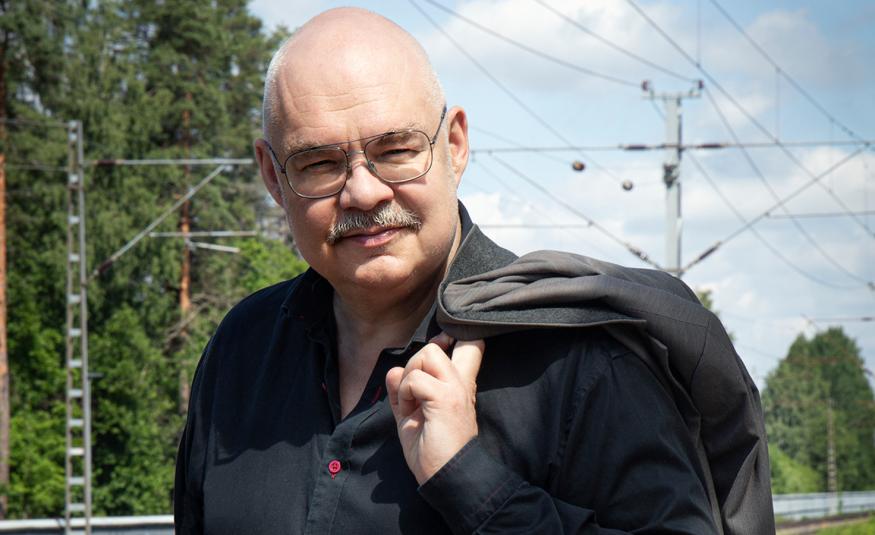 Kuvassa on Mikko Roiha puolivartalokuvassa ja hänellä on musta paita ja musta takki vasemmalla olkapäällään.  Taustalla näkyy metssä ja rautatien sähköjohtoja.