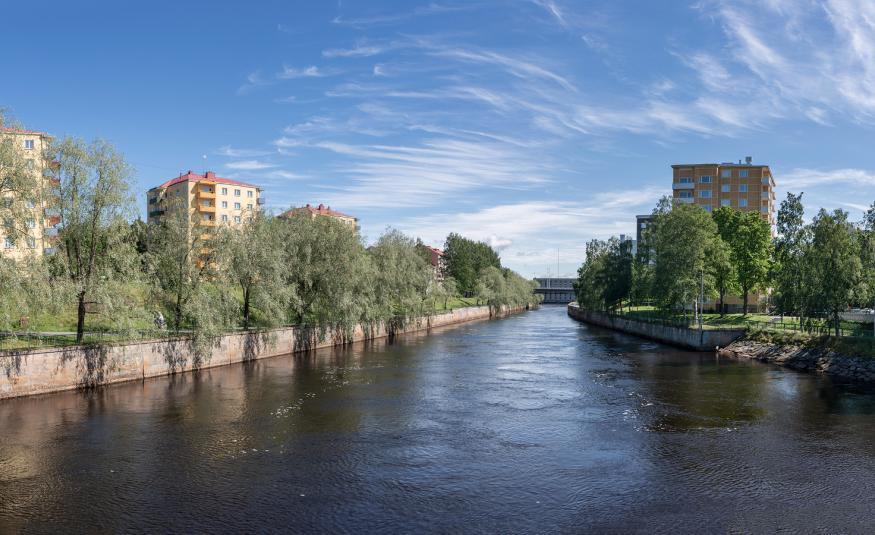Kuvassa on keskellä joki ja sen molemmin puolin kerrostaloja, joiden edessä on lehtipuita. Takana on sinistä taivasta.