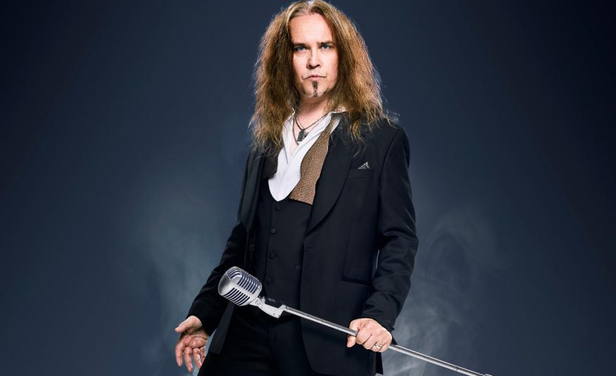 Kuvassa on keskellä on Jarkko Ahola seisomassa ja hänellä musta takki sekä valkoiset kaulukset. Hänellä on jalallinen mikrofoni oikeassa kädessään. Tausta on tumma.