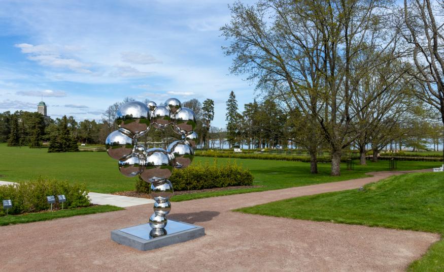  Kuvassa on ruskealla hiekalla teräksinen veistos, jossa on jalustan päällä palloja. Taustalla on vihreää nurmea ja takana metsää. 