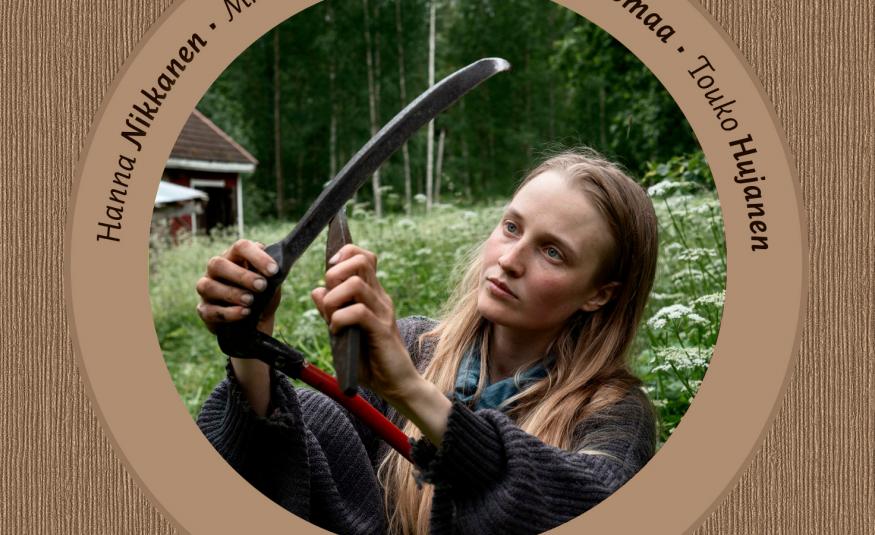 Kuvassa on keskellä ympyrä ja sen sisällä on kuva nuoresta naisesta, joka terottaa viikatetta. Taustalla on vihreää niittyä. Tausta on ruskea ja ympyrän kaarella on tekstiä.