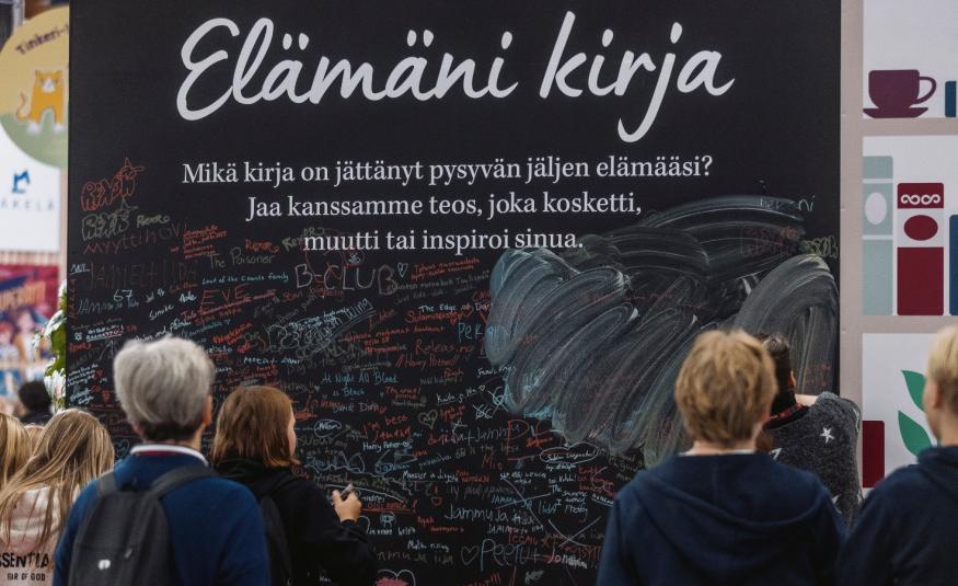Kuvassa on seinämä, jossa lukee kirjoituskirjaimin Elämäni kirja ja sen alla pientä tekstiä mustalla pohjalla. Edessä on ihmisiä katselemassa seinää.