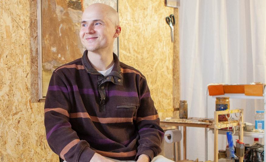 Kuvassa on Kristo Saarikoski ja hänellä on ruskearaitainen pusero päällään. Taustalla on vaaleaa seinää taideteos sekä oikealla vaaleita huonekaluja.  