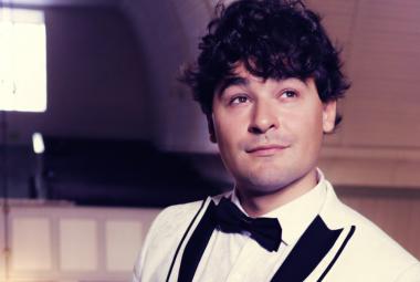 Kuvassa on Amadeus Lundberg puolivartalokuvassa kuvan oikeassa laidassa. Hänellä on valkoinen takki, jossa on kauluksessa mustat kantit. Hänellä on musta rusertti ja valkoinen paita.