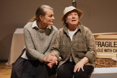 Kuvassa ovat istumassa vierekkäin käsi kädessä vasemmalla Jonna Järnefelt ja vieressään Eija Ahvo.  Heillä on ruskea harmahtavat vaatteet ja Ahvolla on valkoinen hattu päässään. Taustalla on ruskeaa seinää.  