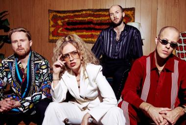 Kuvassa on Haloo Helsingin neljä jäsentä, joista kolme istuu ja yksi seisoo takana. Vasemmalla on mieshenkilökirjavassa takissa ja hänen oikealla puolellaan on nainen valkoisessa housupuvussa. Oikealla istuu mies punaisessa paidassa. Näiden kahden välissä takana seisoo mies, jolla on tumman sininen paita päällään. He ovat ruskeasävyisessä huoneessa. 