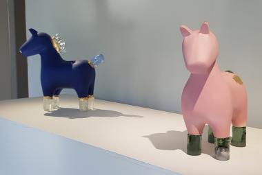 Kuvassa on kaksi lasihevosta hyllyllä. Vasemmalla on sininen hevonen ja oikealla vaalean punainen valkoisella hyllyllä. Tausta on valkoinen.