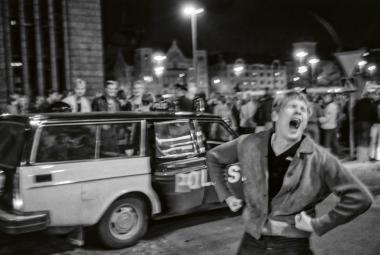 Kuvassa on edessä keskellä nuori mies pusakassa ja hän huutaa. Takana on poliisiauto ja sen takana nuorisojoukkoa Helsingin päärautatieaseman edessä yöllä. Kuva on musta-valkoinen.