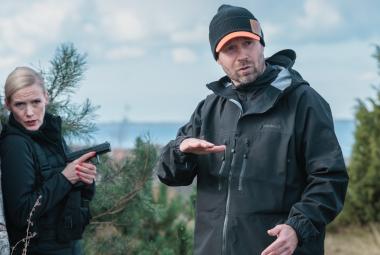 Kuvassa ovat vasemmalla harmaaseen kiveen nojaamassa Laura Birn mustassa takissa pistooli kädessään ll ja oikealla Jyri Kähkönen tummassa anorakissa. Taustalla on  laajaa vihreää aavaa tunturimaisemaa. 