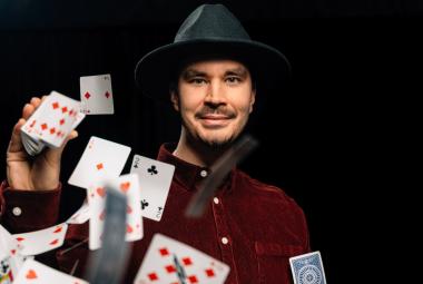 Kuvassa on Miika Storm mustassa lierihatussa ja puserossa puolivartalokuvassa. Hänen käsistään leviää vasemmalle pelikortteja. Tausta on tumma.