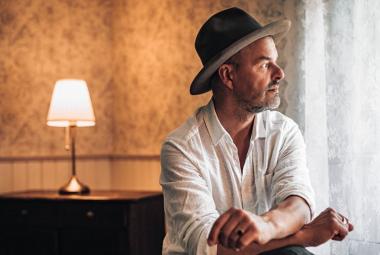 Kuvassa on oikealla Tuure Kilpeläinen istumassa ja hänellä on valkoinen paita ja harmaa hattu. Hän katsoo oikealle ja on sivuttain. Taustalla näkyy vaalean ruskeaa seinää, jossa on kulma. Seinän vieressä on ruskea matala hyllykkö ja sen päällä on pöytälamppu valaistuna.