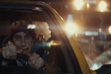 Kuvassa on vasemmalla nuori mies ajamassa autoa yöllä ja autoa näkyy puoliksi. Taustalla näkyy valoja öisellä tiellä ja kuorma-auton valot.