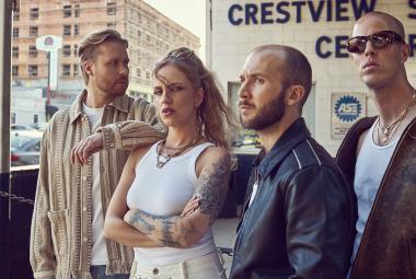 Kuvassa on Haloo Helsingin jäsenet seisomassa tilassa, jossa lukee seinässä Crestview Center. Vasemmalla on mies vaalean ruskeassa villatakissa vieressäänElli valkoisessa hihattomassa t-paidassa. Oikealla on kaksi miestä ja heillä on tummat takit päällään. Lasi-ikkunan takaa näkyy vaalean ruskea iso kerrostalo.