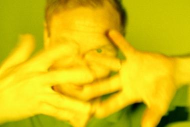 Kuvassa on Ismo Alangon kasvot ja ne ovat edessä olevien käsien takana. Sormet on haristettu. Kuvan sävy on vihertävä.