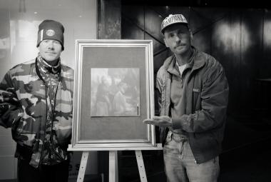 Kuvassa ovat vasemmalla VilleGalle ja oikealla Jare. Heidän välissään on raamitettu taulu varustettuna kolmella jalalla. Kuva on musta-valkoinen.