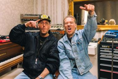 Kuvassa ovat JVG jäsenet ja vasemmalla Ville Gallella on musta nahkatakki ja Jarella vaalean sininen farkkupuku sekä vasen käsi ylhäällä. Taustalla on on soittimia ja äänitysstudio.