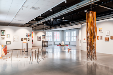 Kuvassa on vaalean harmaat seinät taidenäyttelytilassa ja siinä on eri kokoisia taideteoksia seinillä ja takana yksi pylväs.