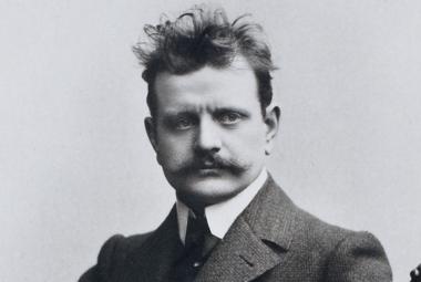 Kuvassa on  Jean Sibelius puolivartalokuvassa ja hänellä on hiukset päälaella pörhöllään ja paksu takki päällään sekä valkoiset kaulukset. Kuva on musta-valkoinen.