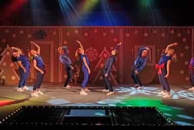 Kuvassa on kahdeksan henkilöä sinisissä asuissa taipuneina taaksepäin toinen käsi ojennettuna ylös. Taustalla on ruskeaa seinämää, jossa on ruskehtavia joulukuusia.