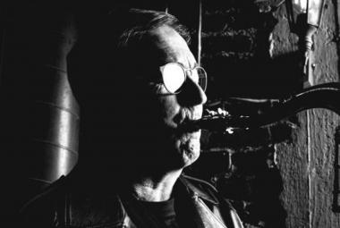 Kuvassa on Jussi Saksa soittamassa saksofonia puolivartalokuvassa. Kuva on tumma ja silmälasit kiiltävät ja kasvot näkyvät osittain.