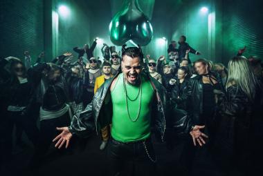 Kuvassa on keskellä Käärijä vihreässä puserossa ja mustassa nahkatakissa. Taustalla on joukko ihmisiä ja valoja ohjattuna heihin. 