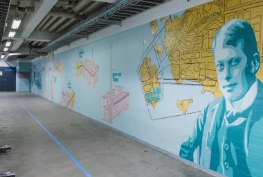 Kuvassa on käytävä, jossa on oikean puoleinen seinä ja sen pohja on vaalean sininen.  Seinällä on karttapohjamaisia kuvioita ja oikealla keltainen niemi.  Lähinnä oikealla on iso miehen rintakuva vaalean sinisessä takissa.