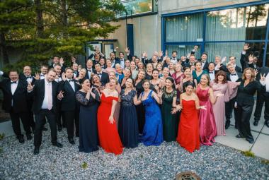 Kuvassa on koko kansallisoopperan kuoro seisomassa pihalla. Vasemmalla ja takana ovat miehet musta-valkoisissa puvuissa. Edessä ovat naiset pitkissä, eri värisissä pitkissä iltapuvuissa. 