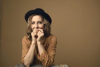 Kuvassa on Mariska puolivartalokuvassa ja hänellä on musta hattu päässään sekä kellertävä, pikkukuvioinen pusero päällään. Hänellä on molemmat kädet kevyesti nyrkissä suun edessä. Tausta on ruskehtavaa seinää.