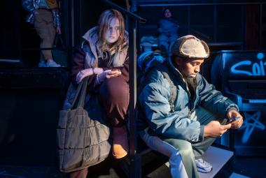 Kuvassa on oikealla nuori mies istumassa karvahatussa ja toppatakissa. Hänen takanaa vasemmalla on nuori nainen, jonka hiukset roikkuva ja hän nojaa vasempaan jalkaansa. Tausta on osittain tumma. 