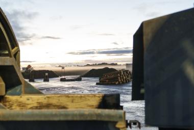 Kuvassa on vasemmalla kellertäviä mrtallirakenteita.  Edessä on vettä ja kauempanapuupino.  Oikealla on tumma, iso  metallinen seinämä.