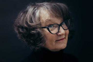 Kuvassa on Monika Fagerholmin kasvokuva ja hänellä on kihartavat tumman harmaat hiukset  sekä mustasankaiset silmälasit. Tausta on musta.