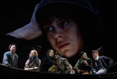 Kuvassa on veneessä kuusi henkilöä tummahkoissa vaatteissa. Taustalla on tummalla taustalla nuoren naisen kasvokuva lierihatussa.