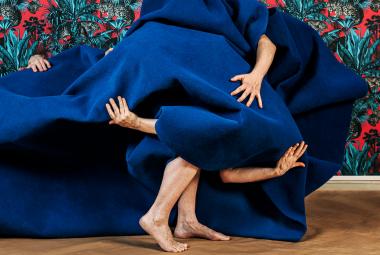 Kuvassa on sinisen kankaan alla henkilöitä ja käsiä näkyy kankaan välistä ja keskellä alhaalla on kävelevät jalat. Taustalla on kukallista puna-turkoosi-mustaa tapettia.