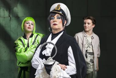 Kuvassa on vasemmalla nuori vaalean vihreässä pusakassa ja hiukset ovat saman väriset. Hänen vieressään on nainen valkoisessa kapteenin hatussa ja mustassa liivissä sekä valkoisessa paidassa. Oikealla on nuori mies harmaassa pusakassa ja lippalakissa.