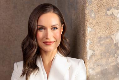 Kuvassa on Sanna Marin puolivartalokuvassa ja hänellä on valkoinen jakku päällään, jossa on v-aukko. Hänellä on pitkät tummat hiukset. Tausta on vaalean ruskeaa kiviseinää.